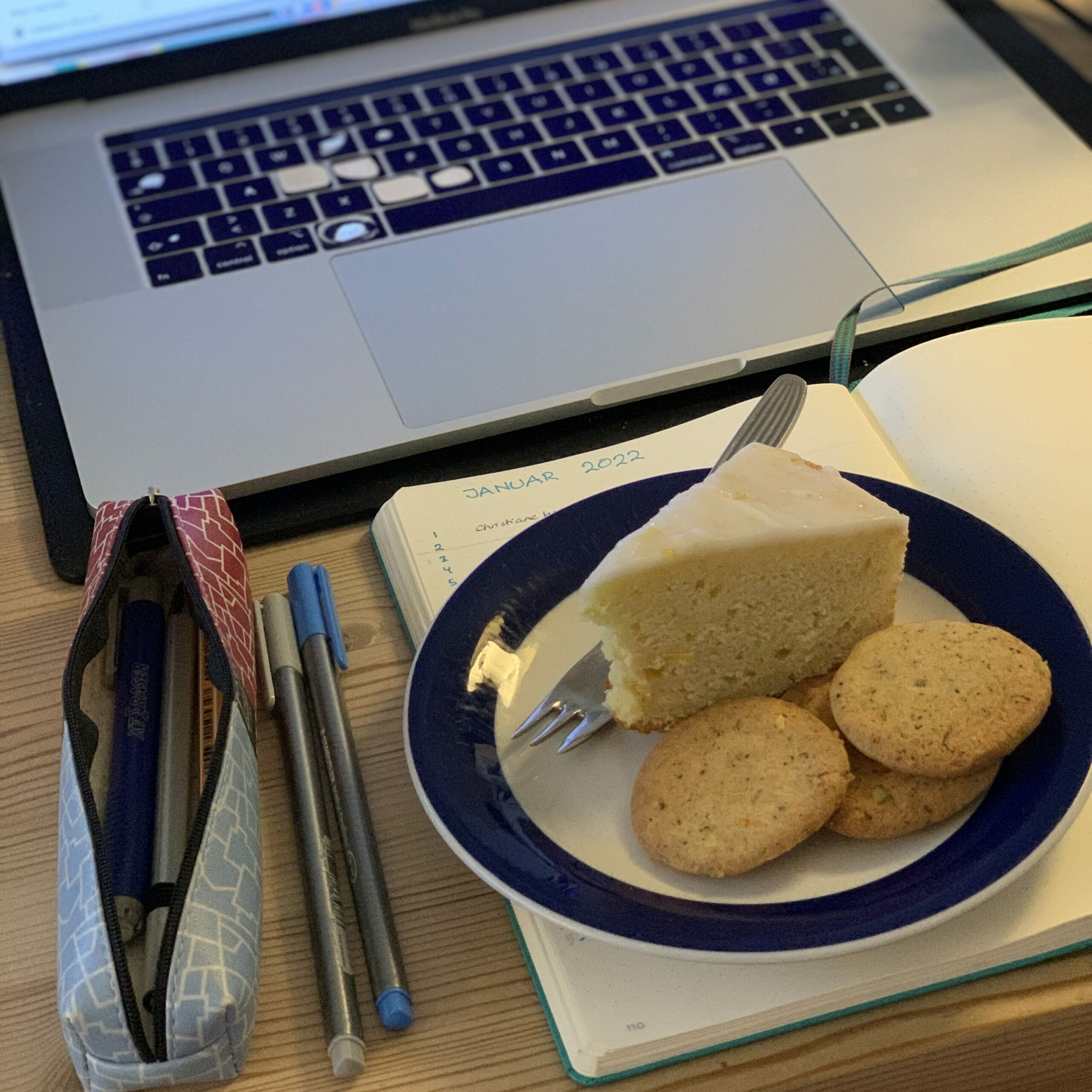 En arbejdsplads med åben laptop, notesblok og pennalhus - og en tallerken med et bastant stykke citronkage og tre småkager.