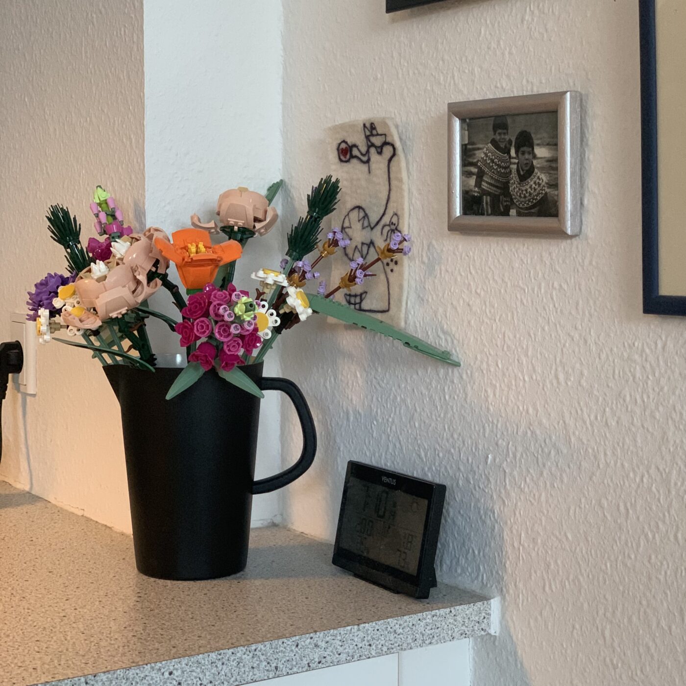 Stilleben med en buket blomster (bygget af legoklodser) på bordet, og forskellige billeder på væggen. Det ene billede er et sort/hvidt foto af et par piger i grønlandsk nationaldragt.
