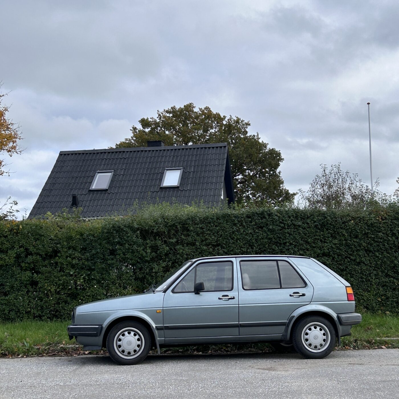 En gammel VolksWagen parkeret foran en grund med ligusterhæk. Bag hækken ses et sort tag med to velux-vinduer.